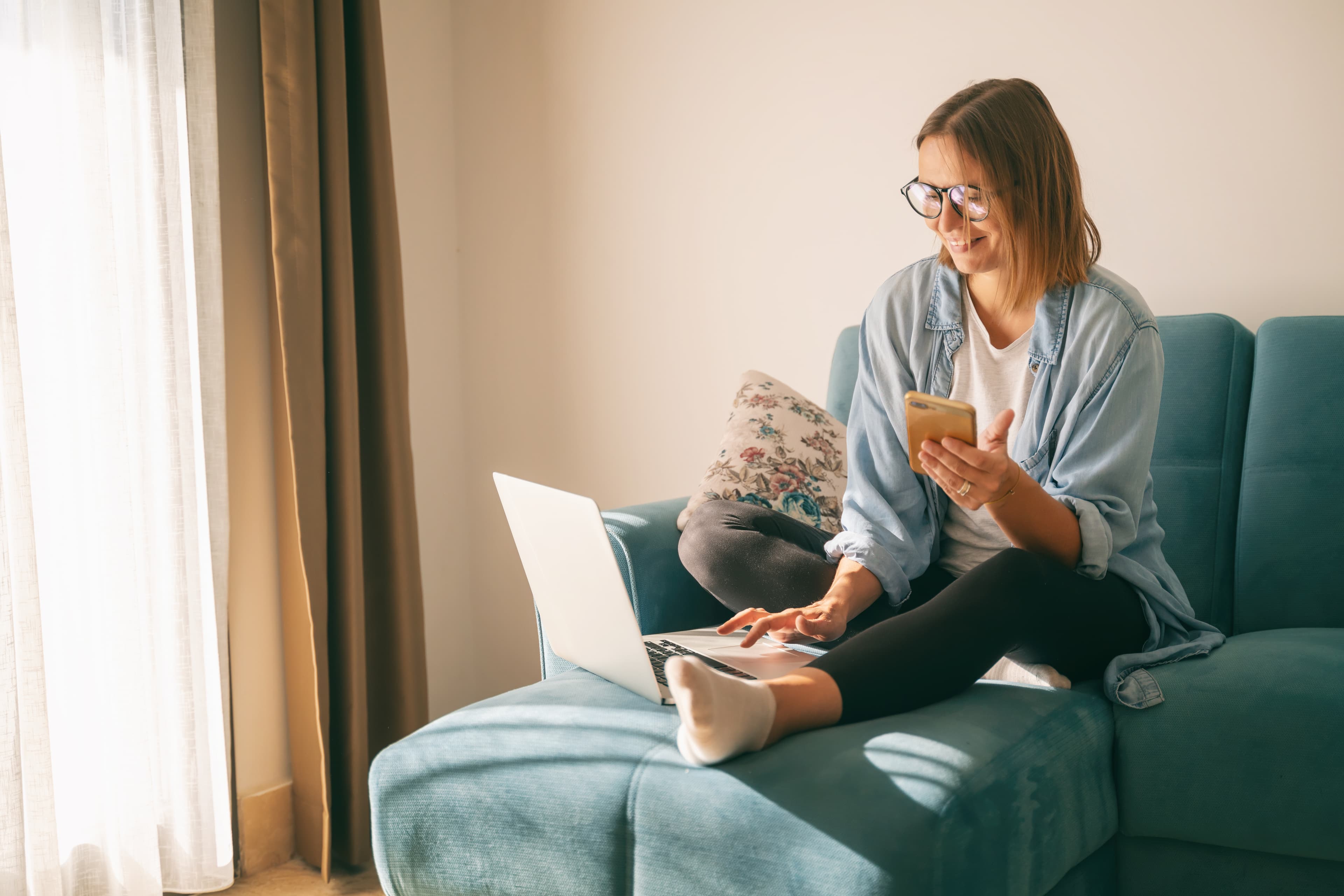 Young mature attractive woman sitting on a blue sofa in the living room at home with a smartphone in her hands in front of a laptop, online shopping.
