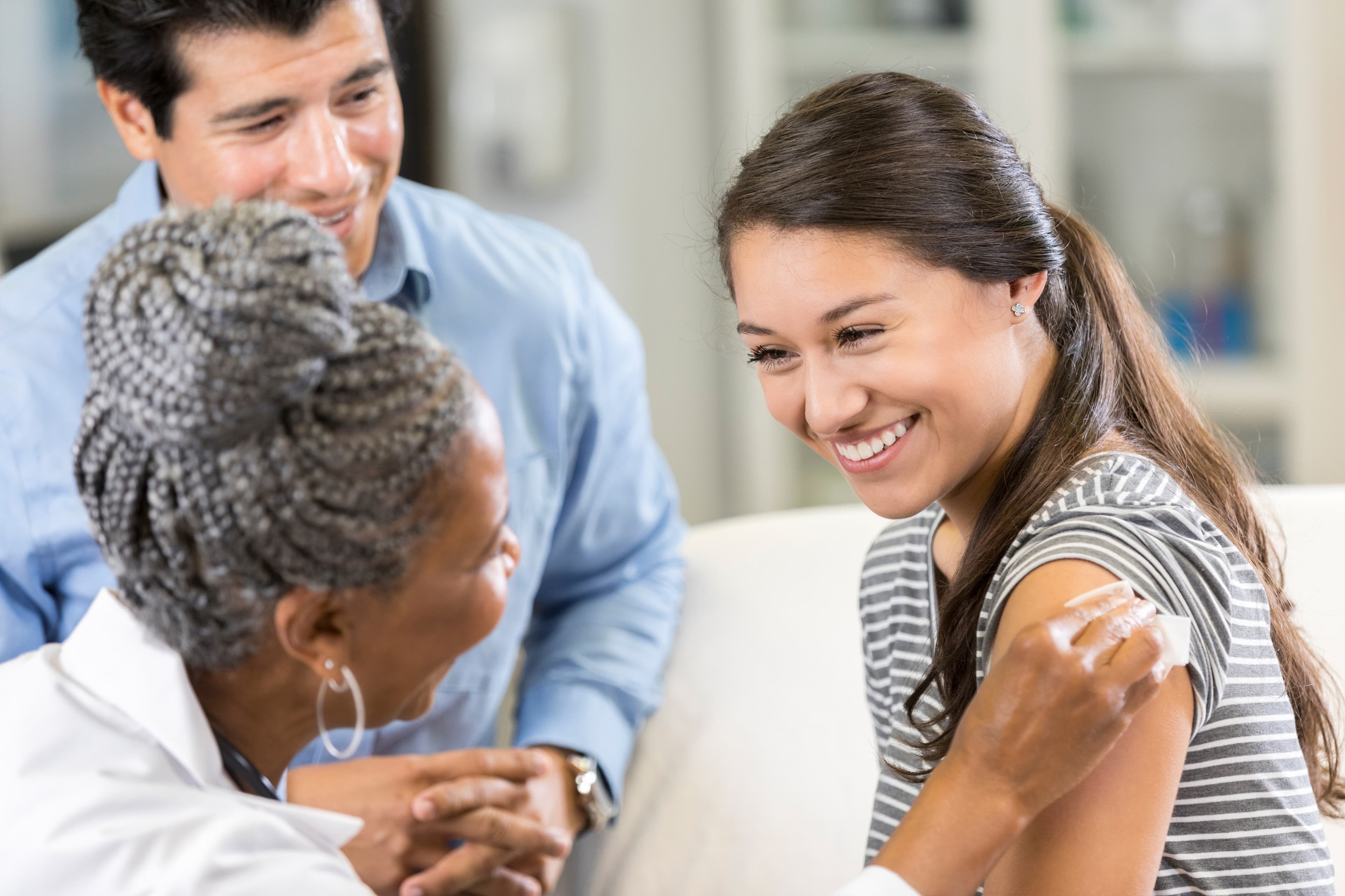 Cheerful teenage girl receives a vaccine from a senior female healthcare professional. The girl's father is sitting next to her.