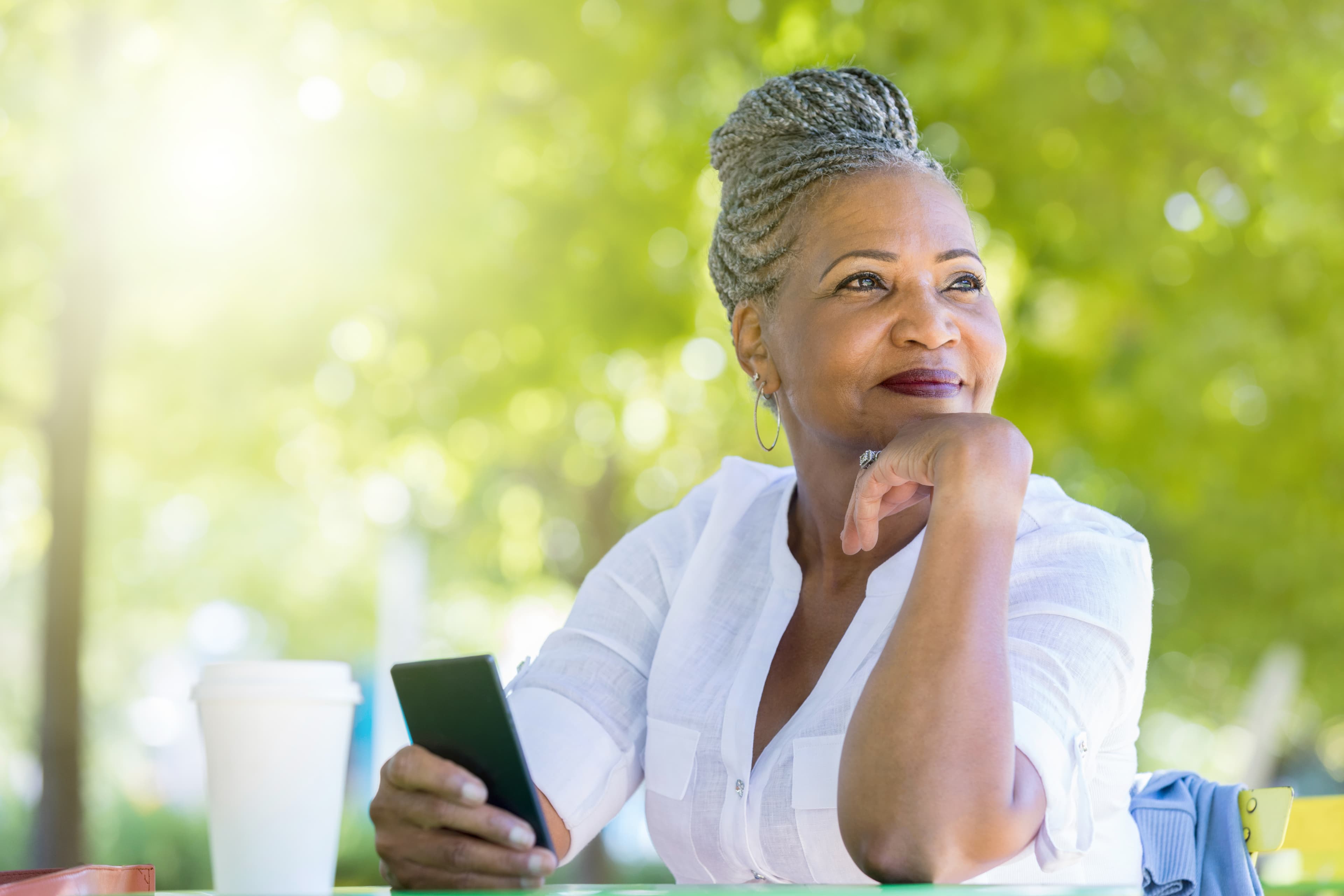 A cheerful senior woman rests her chin in her hand as she looks away.  She is sitting at a table in a public park with coffee and her smart phone and waiting for a friend.