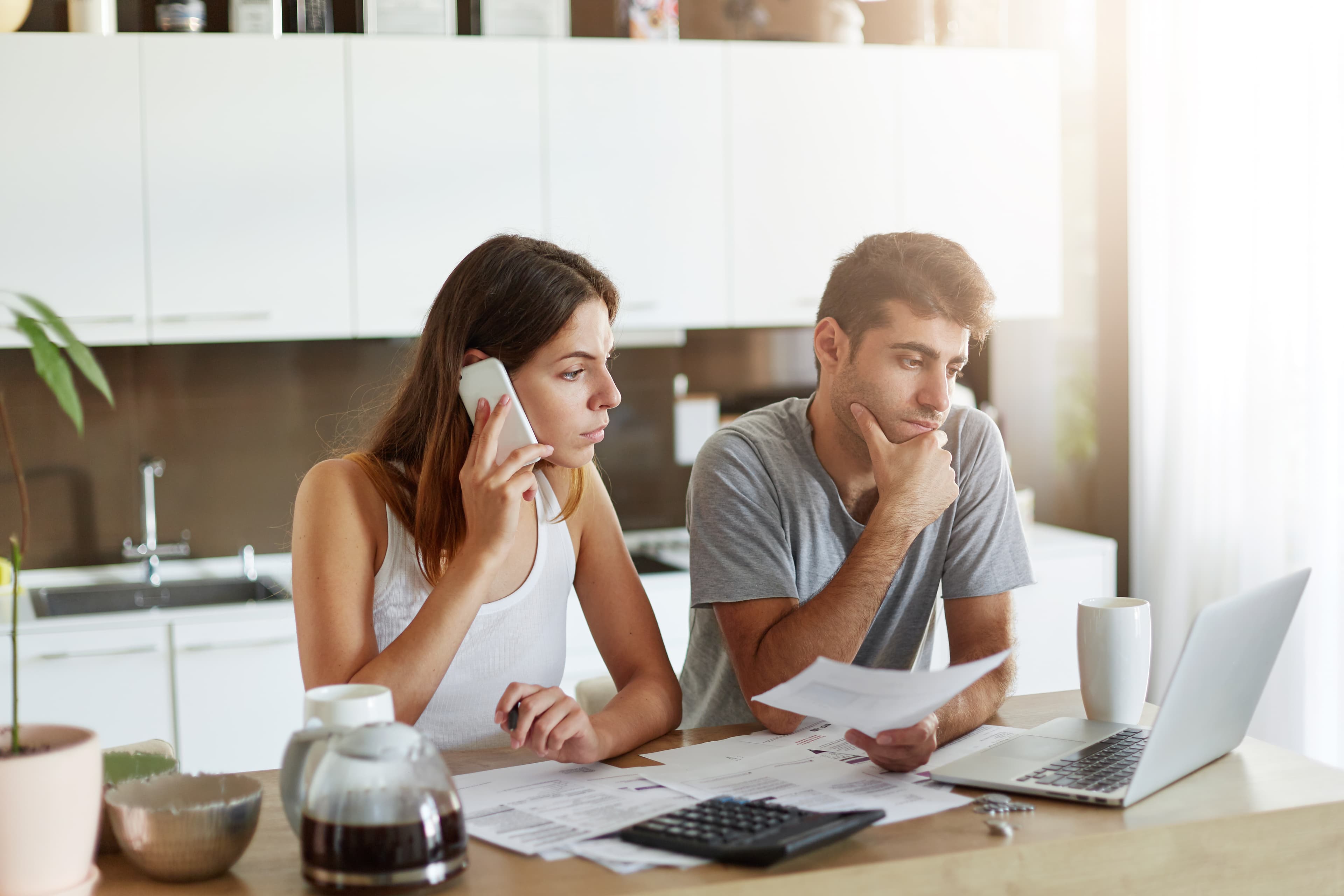 Business couple working on their project, sitting at kitchen table, surrounded with documents, laptop and calculator. Young female freelancer looking attentively into screen of laptop while chatting