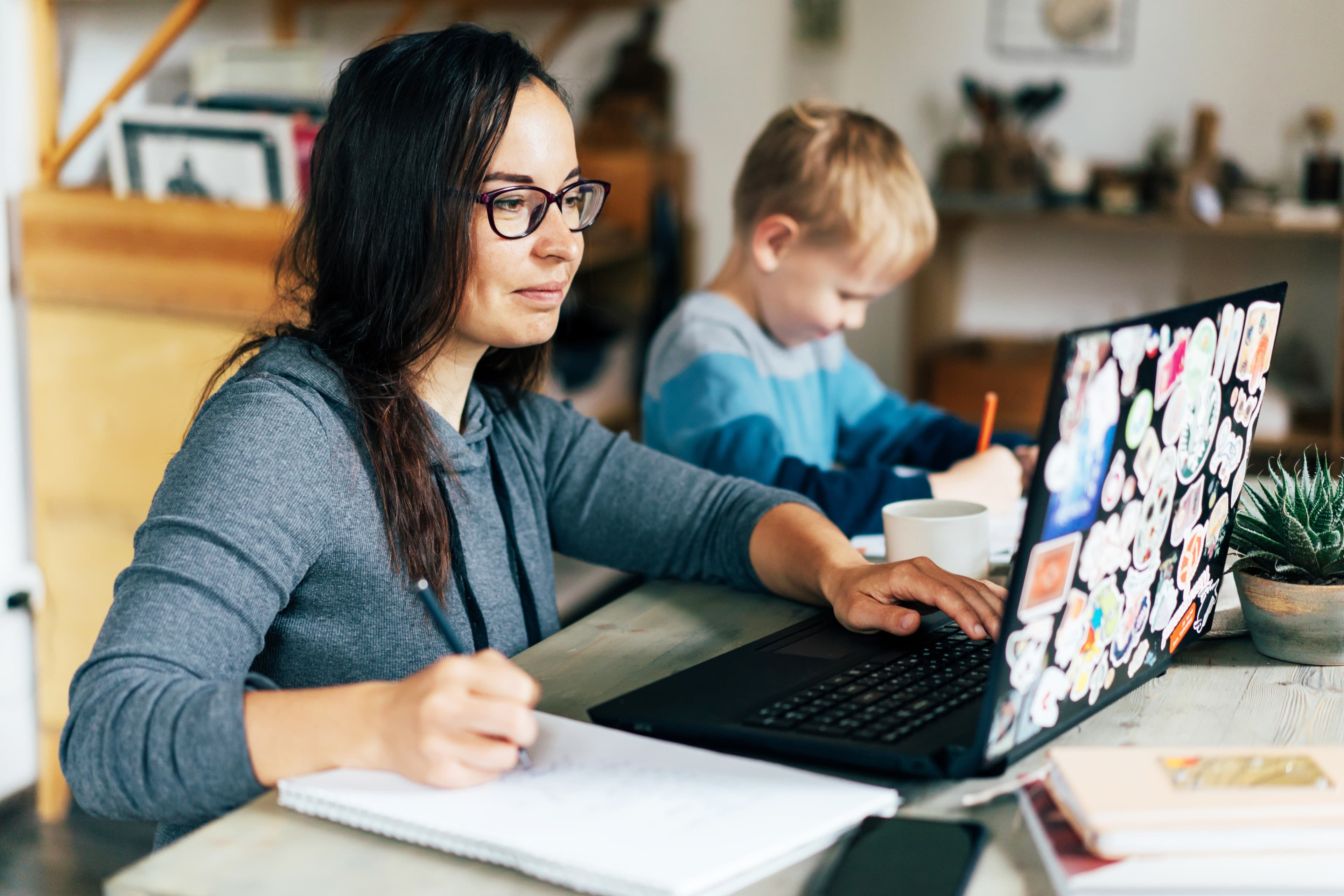 Concept of work from home and home family education. Mom and son are sitting at the desk. Business woman works on the Internet in a laptop, a child writes in a notebook.