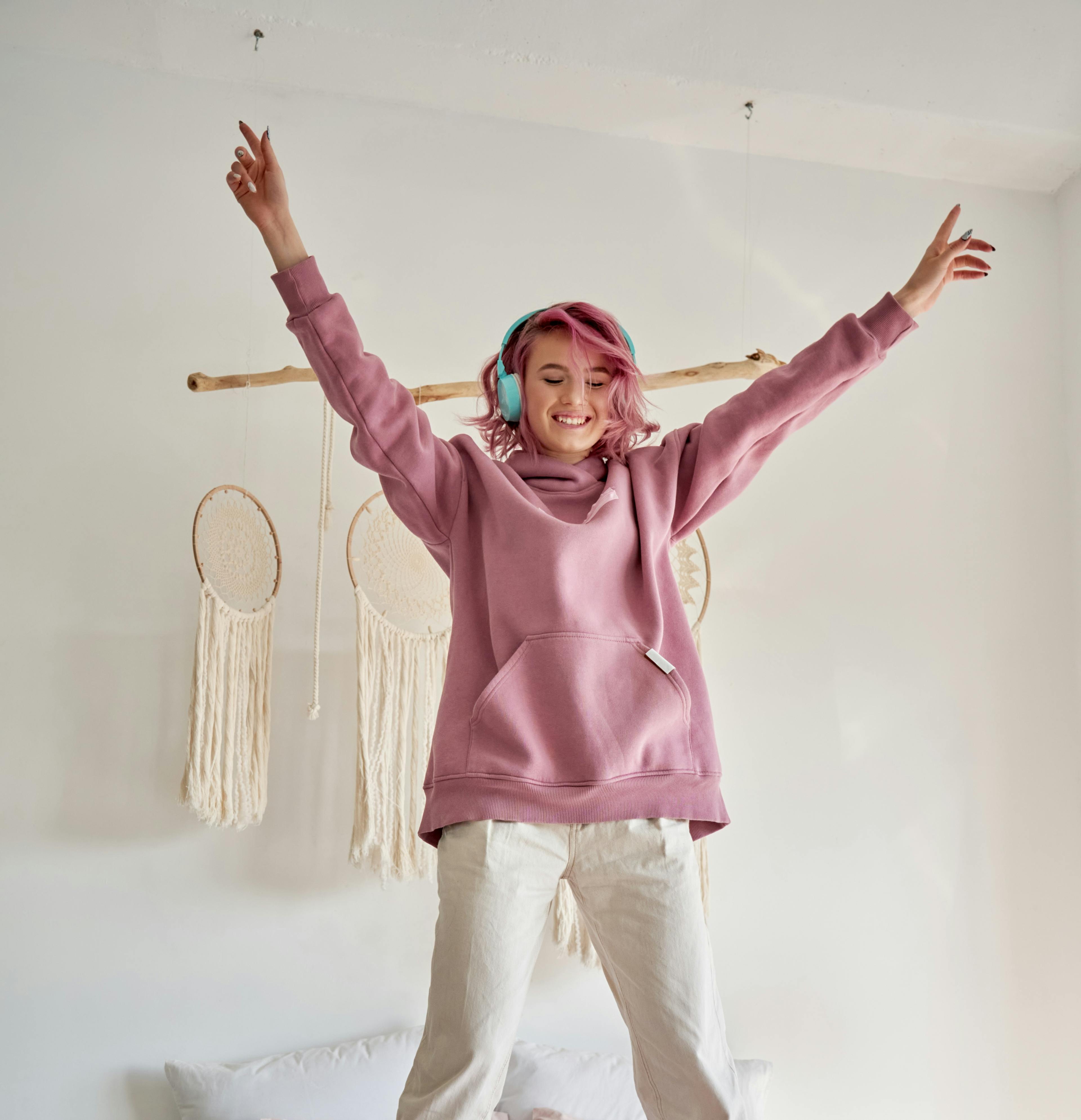 Happy funny funky hipster gen z teen girl with pink hair wear hoodie headphones listening radio pop music playlist podcast, jumping dancing having fun standing on bed in cozy bedroom. Vertical