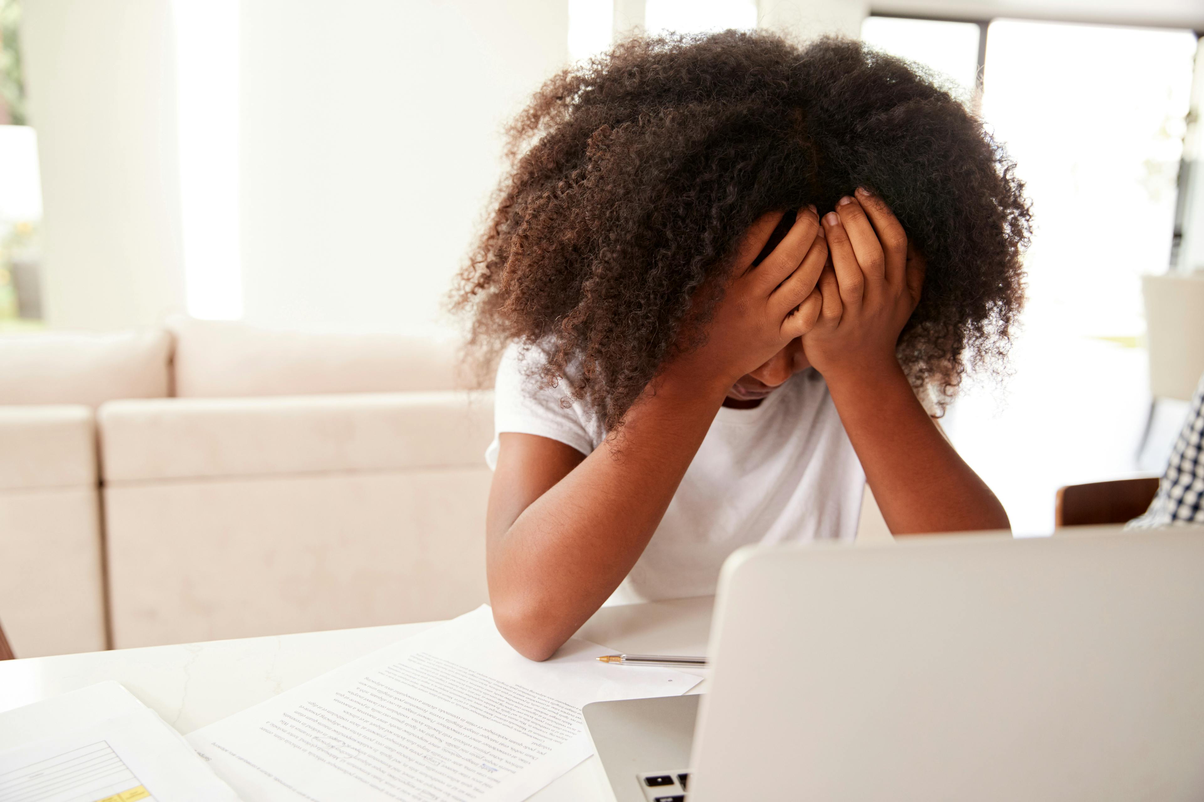 Depressed young teenage black girl with head in hands using laptop computer at home, close up