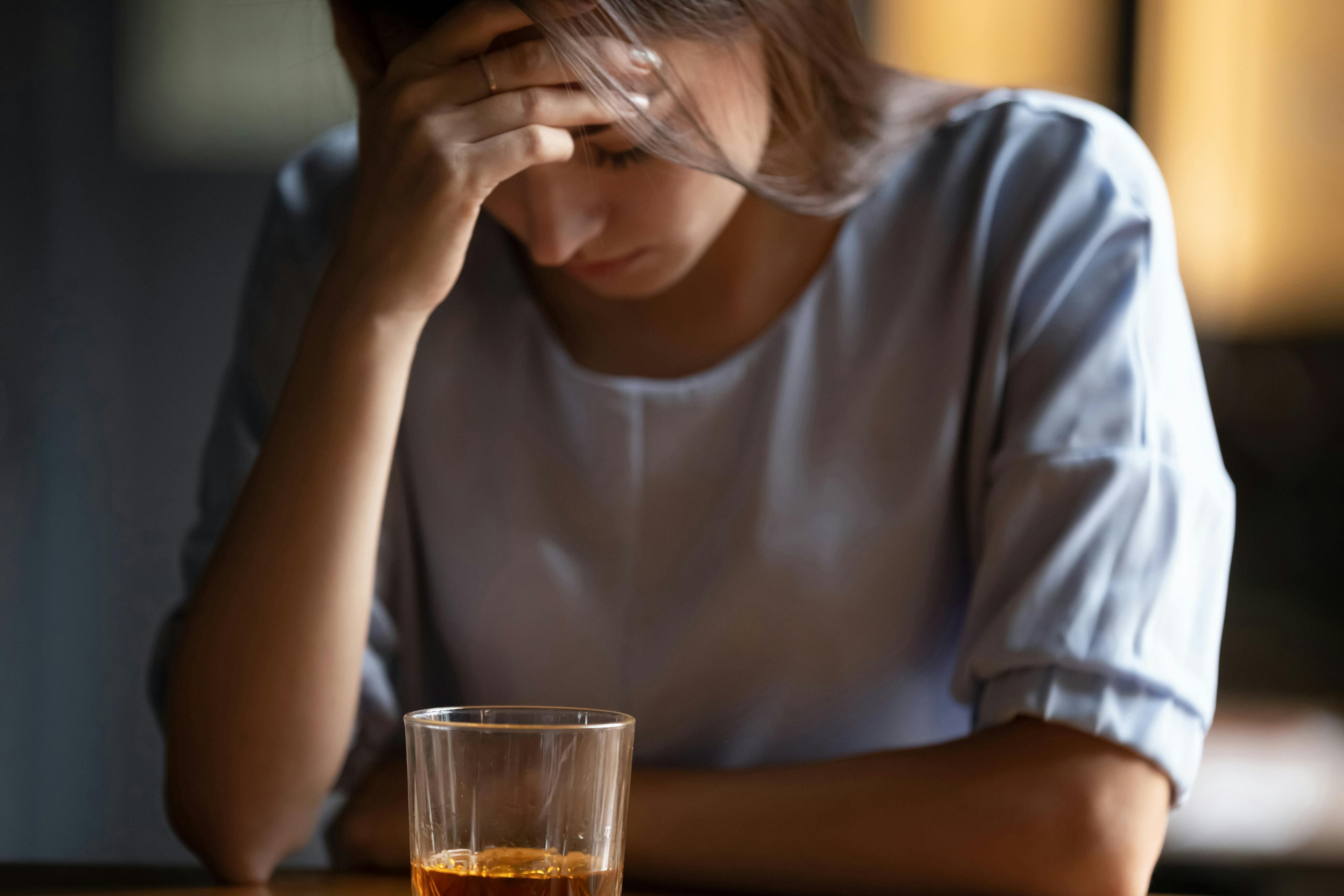 Close up unhappy woman sitting alone with glass of whiskey, touching forehead, thinking about problem at work or relationship problems, feeling desperate and depressed, alcohol addiction concept