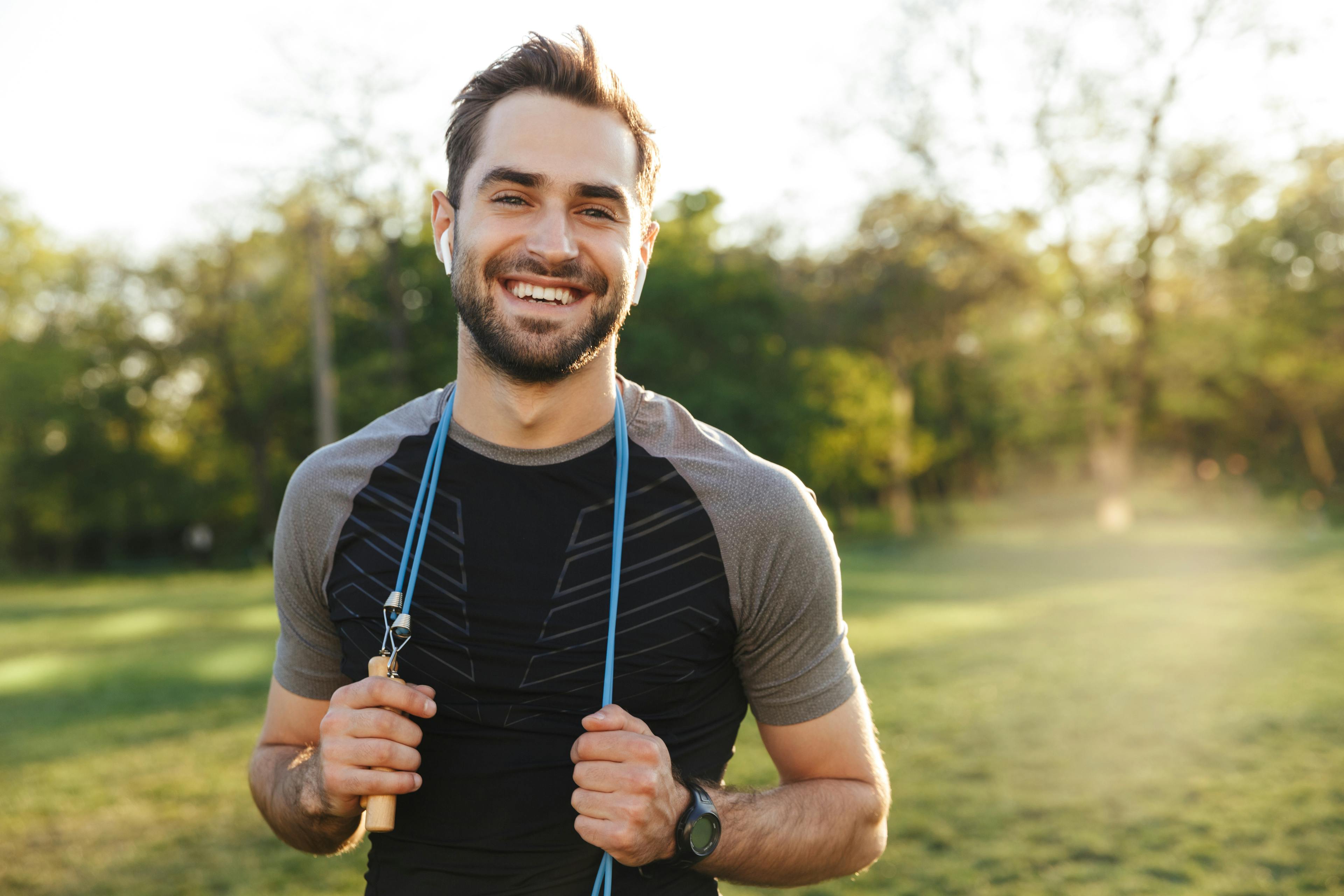 Image of a handsome young strong sports man posing outdoors at the nature park location with skipping rope.