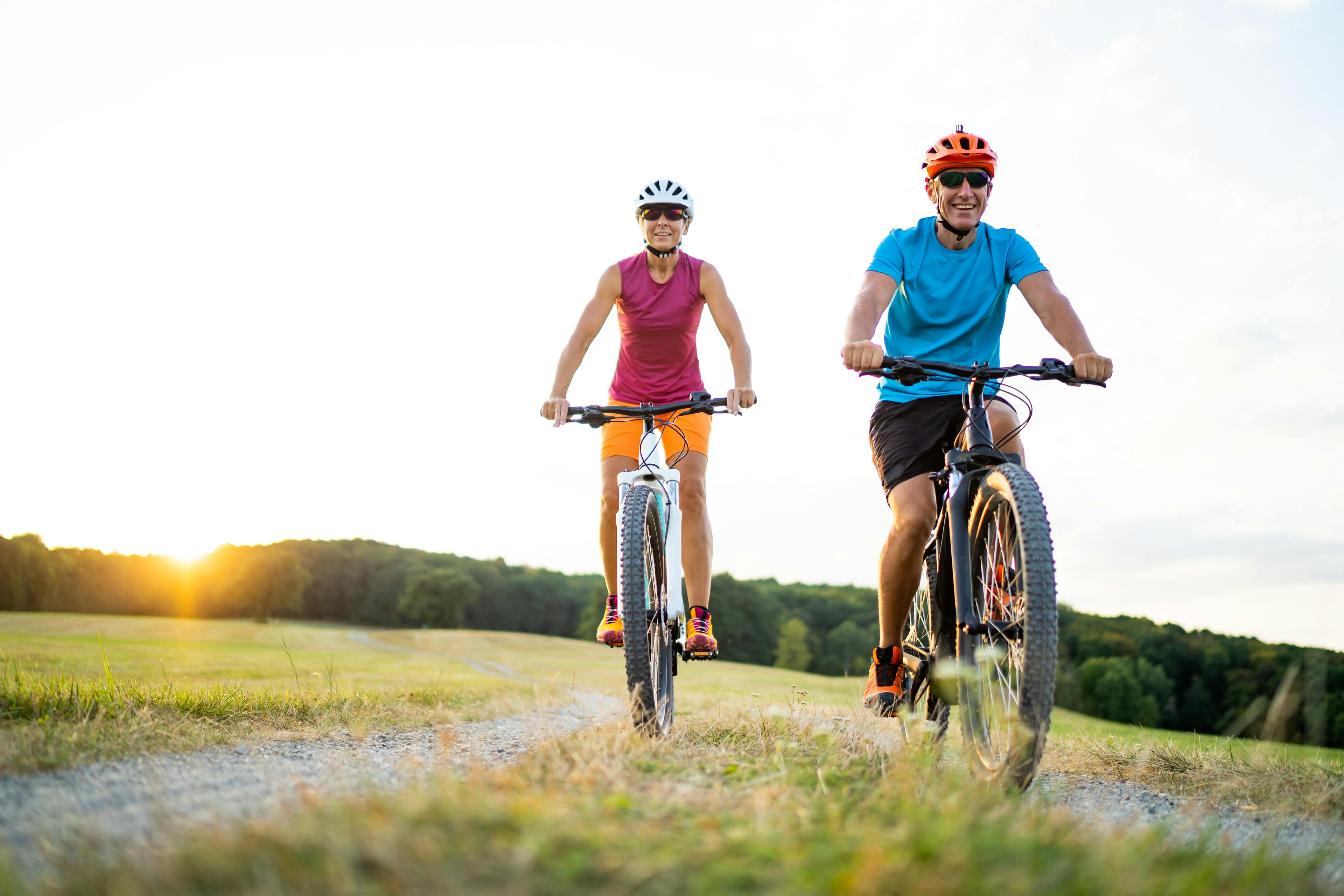 low angle view athletic sporty fit happy smiling  woman and man couple friends in their fourties cycling with their electric mountain bicycles on gravel path through grassland rural landscape on sunny summer afternoon sunset