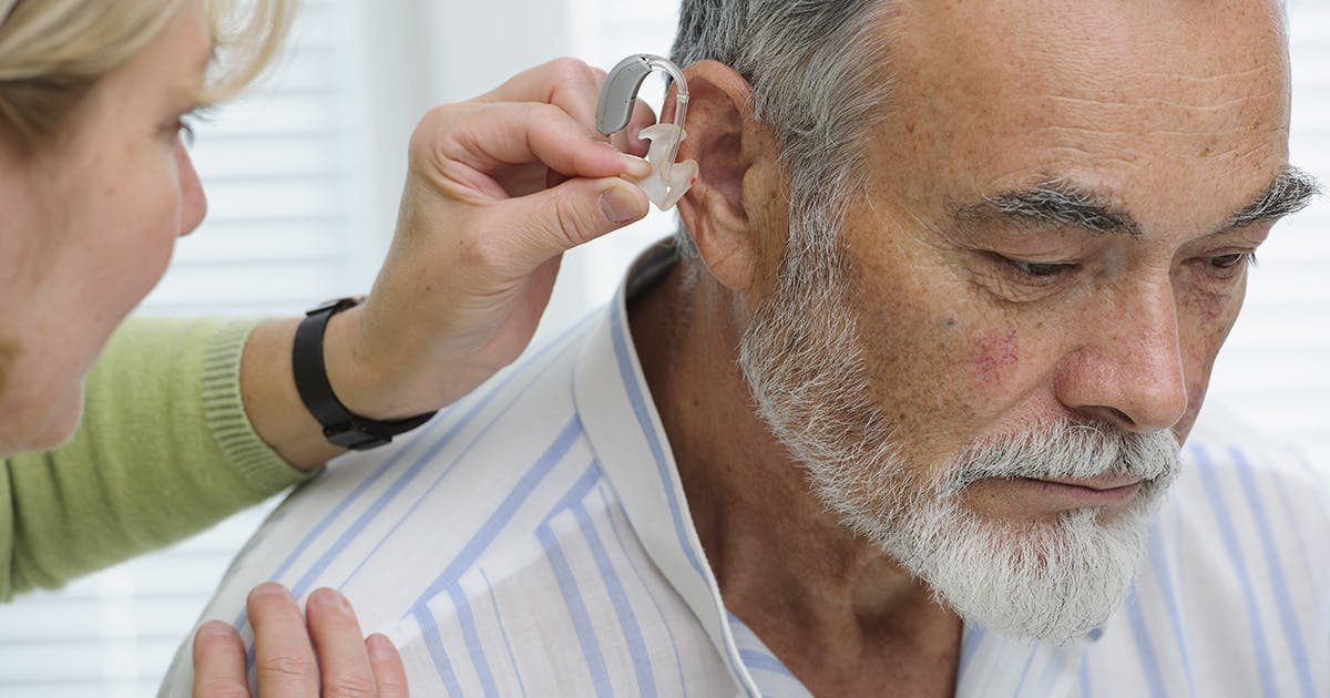 A man with a silver beard and graying hair tilts his head forward as a healthcare professional in a green shirt prepares to fit a hearing aid into his ear.  