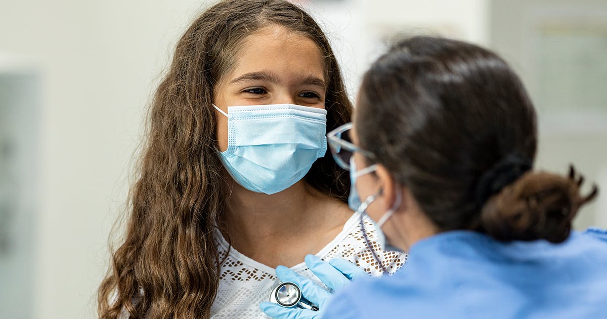 A child wearing a mask is having her heart listened to by a nurse who is also wearing a mask.