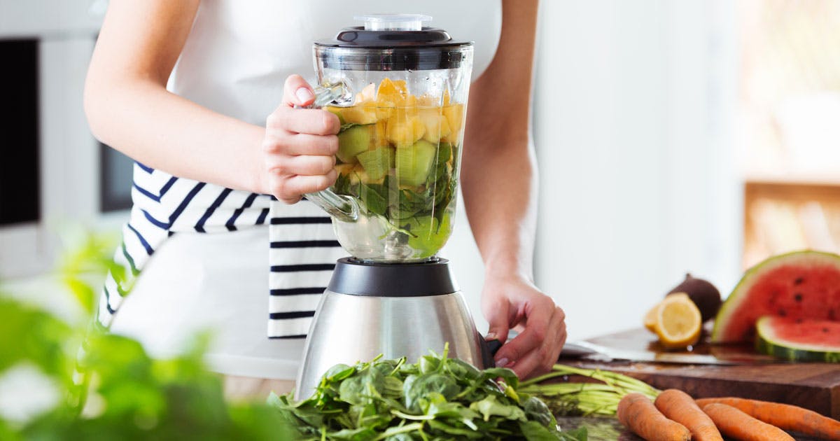 A woman is using a blender in a kitchen to make a fruit and vegetable smoothie.