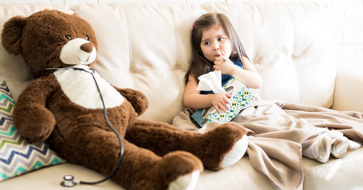 A young girl is sitting on a couch with a thermometer in her mouth and holding a box of tissues.