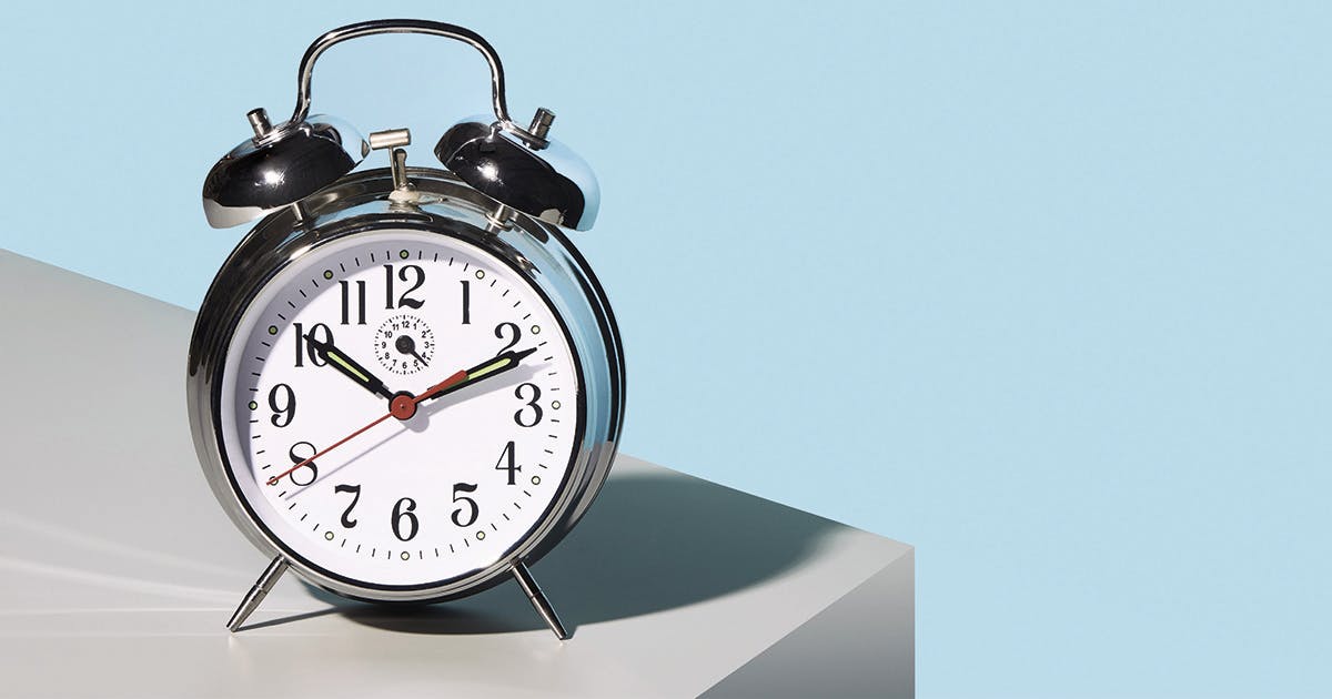 A silver alarm clock sits on a gray table in front of a blue wall
