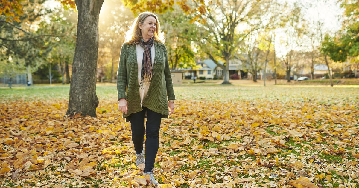 A woman walks through fallen leaves in a park.