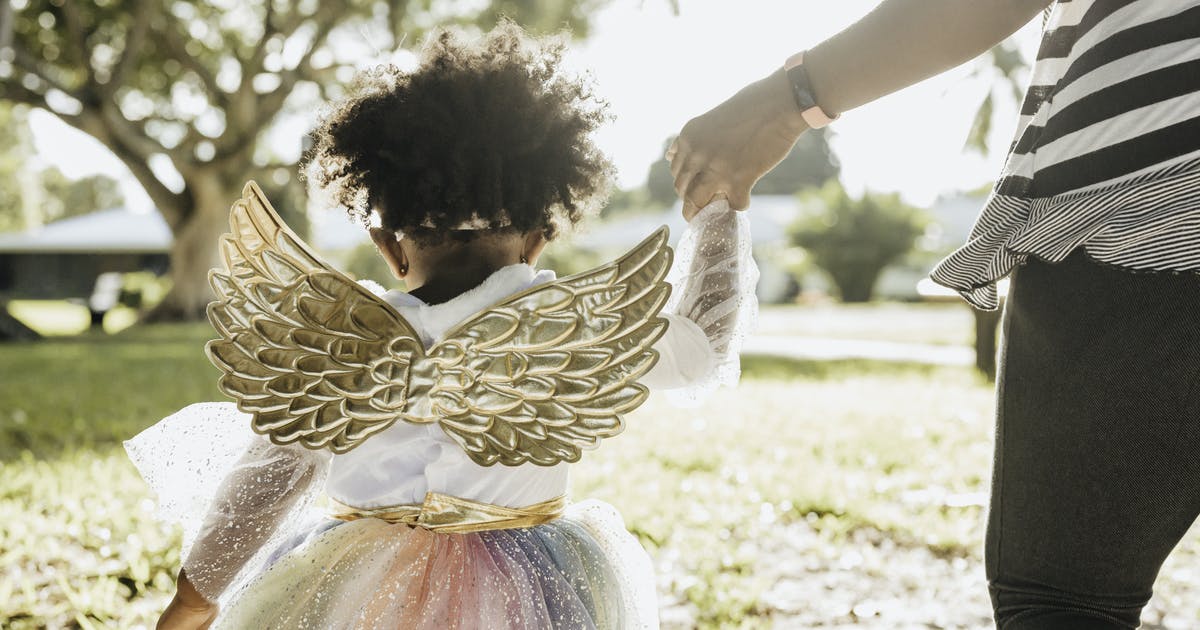 A little girl wearing a tutu and wings holds an adult’s hand.