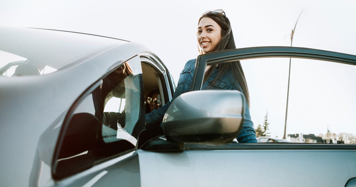 A teen girl with long dark hair wearing a denim jacket gets into the driver’s side of a silver car. 