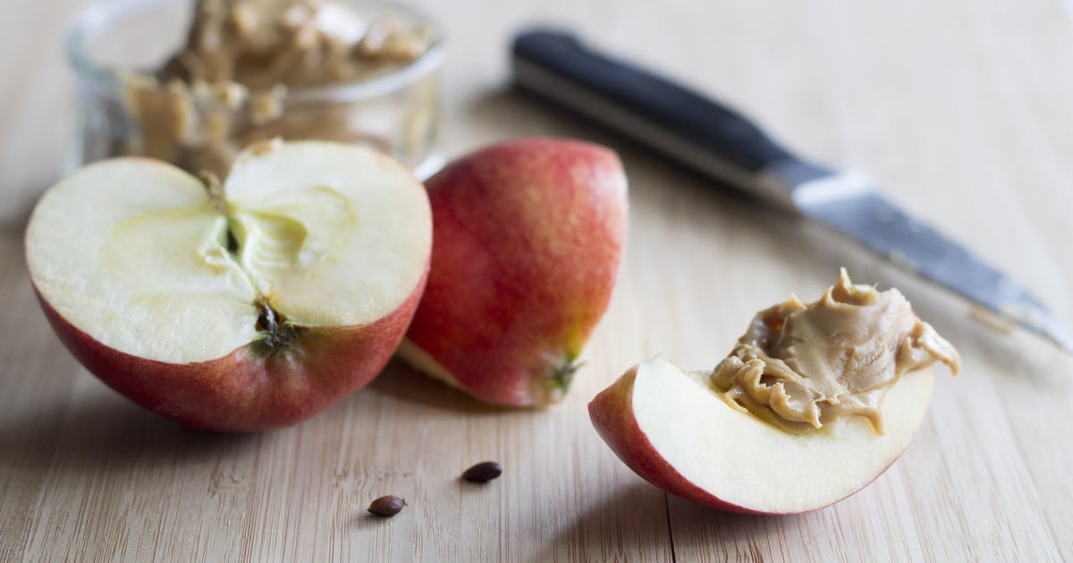 Sliced apples with peanut butter on a countertop. A knife and a bowl of peanut butter sit nearby.