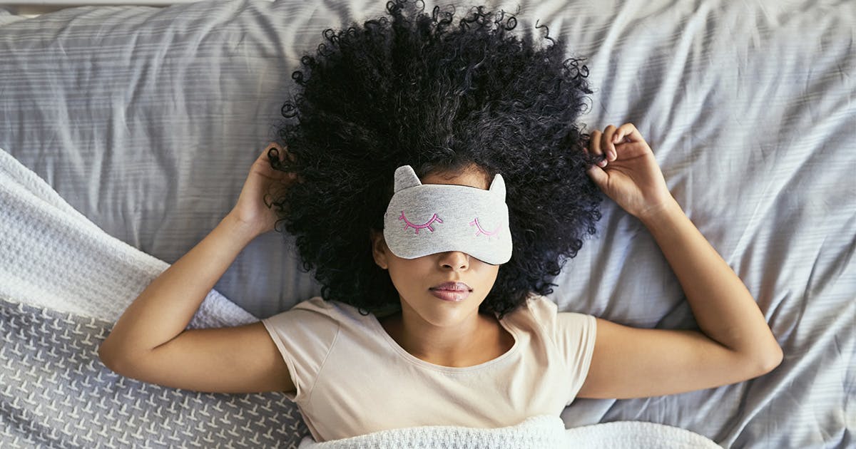 A woman is lying on her back in bed with a sleep mask over her eyes.