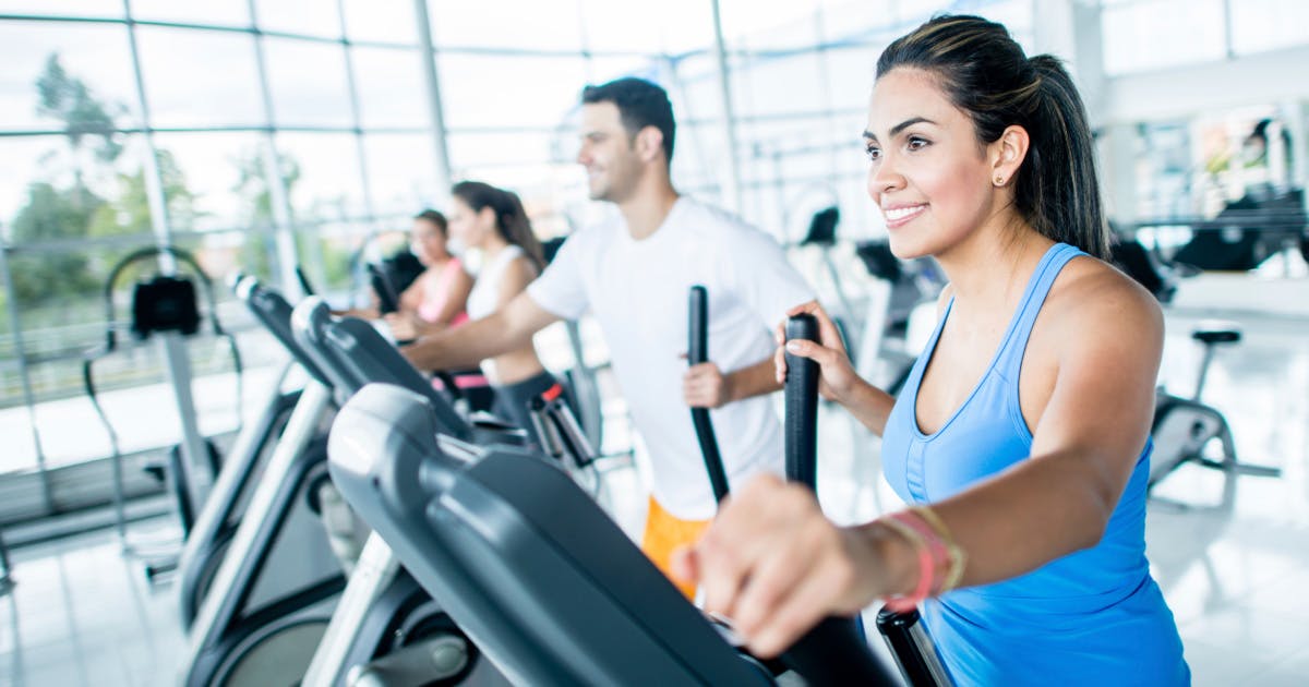 A woman on an elliptical machine with other gym patrons in the background.