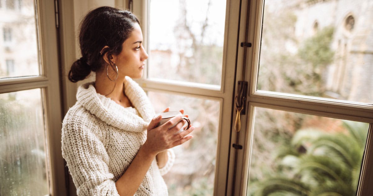 A young woman in a sweater holds a mug and looks out a window.