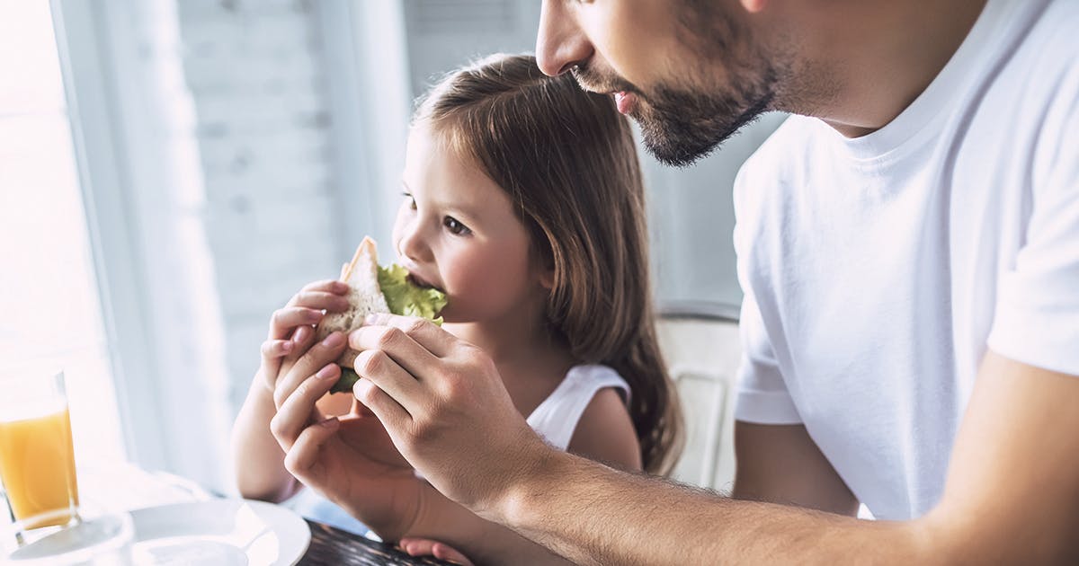 A father helps a child eat sandwich with lettuce.