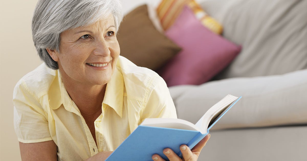 A woman in a yellow shirt with silver hair looks up from her book and smiles.