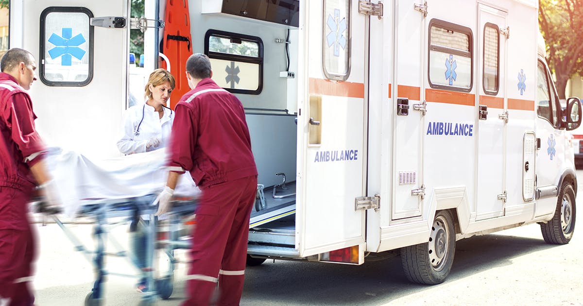 Paramedics in maroon scrubs rush a gurney out of an ambulance as a doctor in a white coat examines the patient.