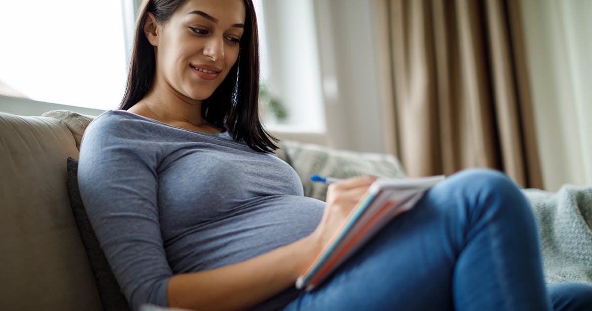 A pregnant woman sits on a couch and writes in a notebook.