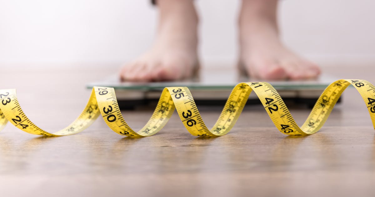 The feet of a woman standing on a scale with a tape measure in front of the scale.