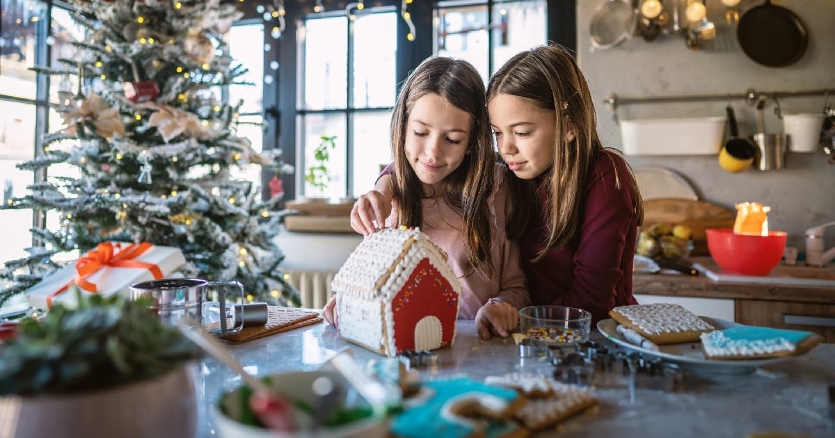 Two young girls decorate a gingerbread house.