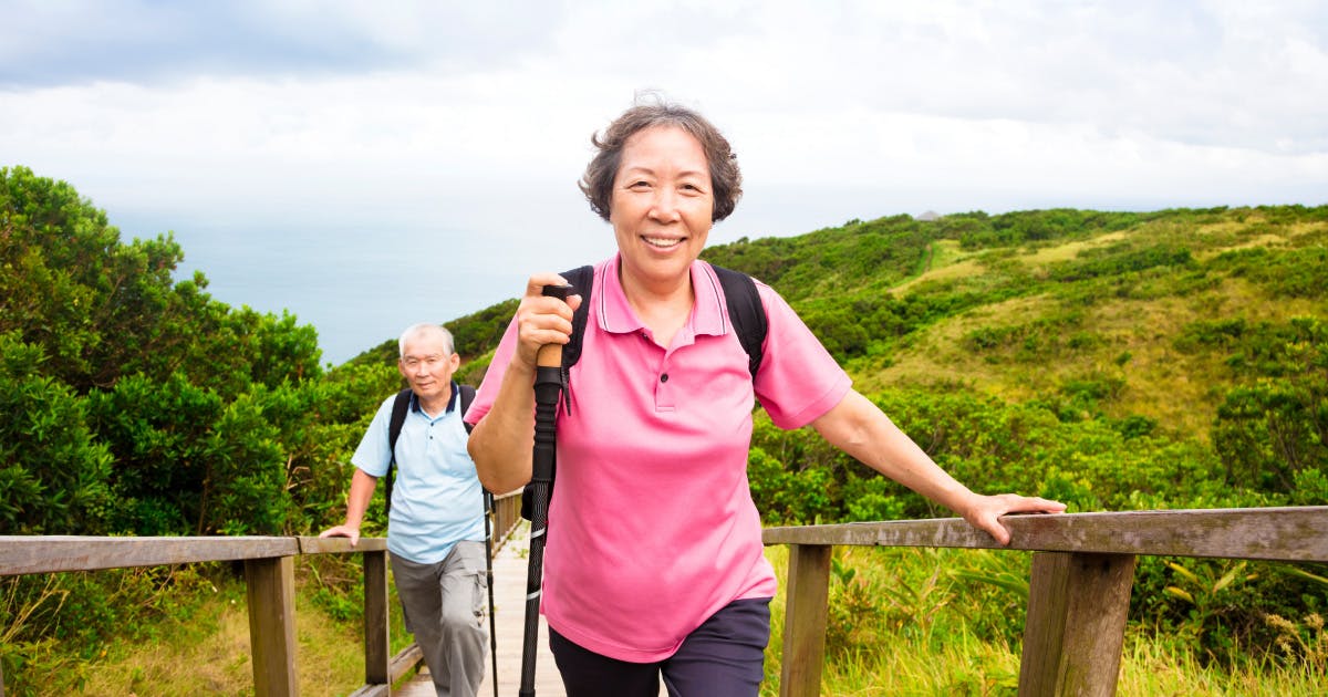 An older couple uses hiking poles on a boardwalk over a marsh.