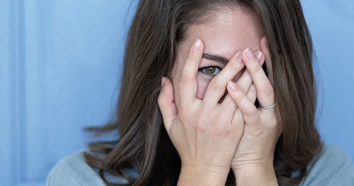 A woman covers her face with her hands. One eye shows through her fingers. 