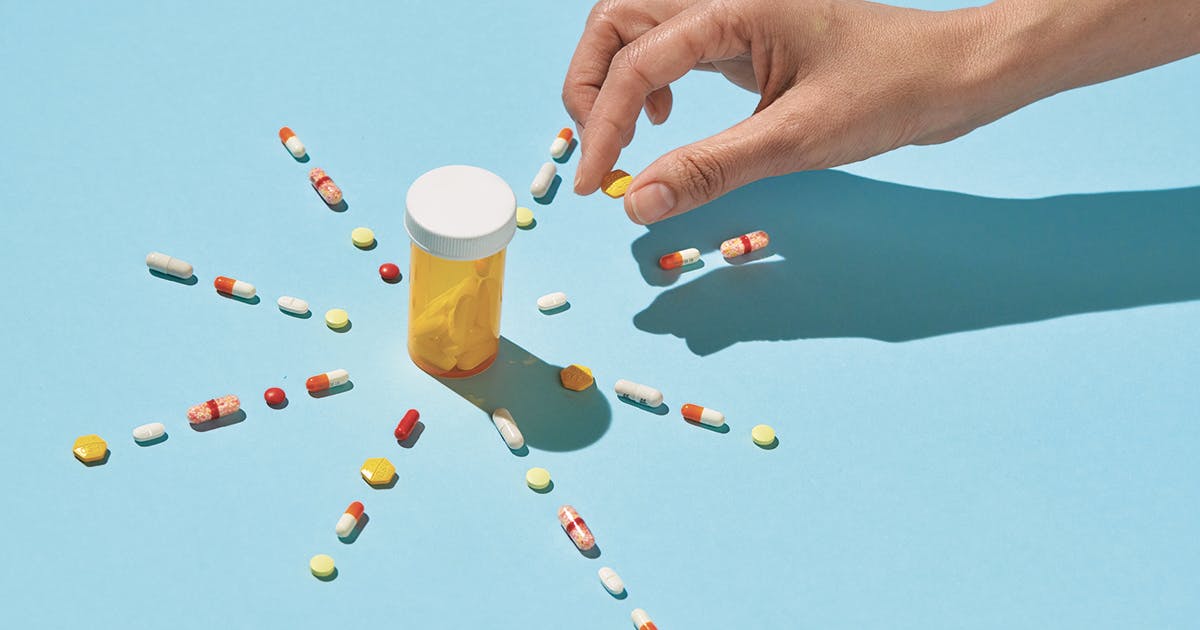 A hand arranges a variety of pills into a star shape around an empty orange pill bottle.