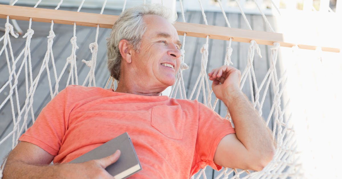 A smiling man reclines on a hammock, holding a book.