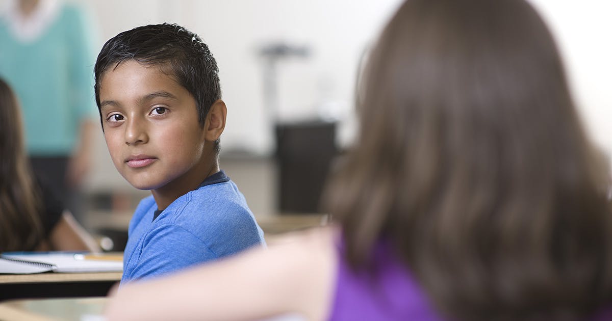 A boy in a blue shirt sits at a desk in a classroom, looking back over his shoulder at a girl in purple. 