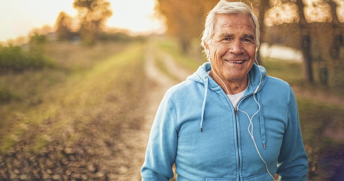 A man with white hair, wearing a turquoise hoodie and white earbuds, walks along an outdoor trail at sunset.
