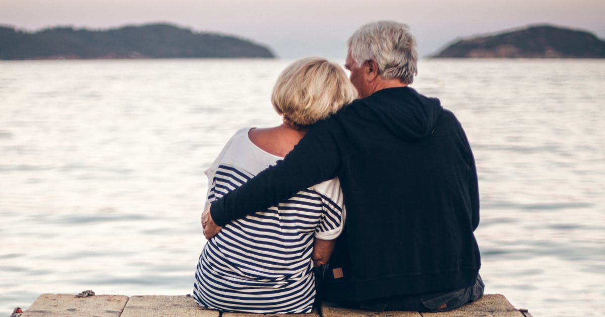An older couple sits on a dock, his arm around her shoulders