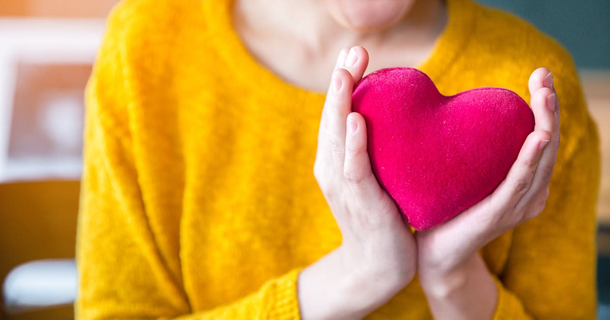  Women in a yellow sweater holds a pink stuffed heart in front of her chest.