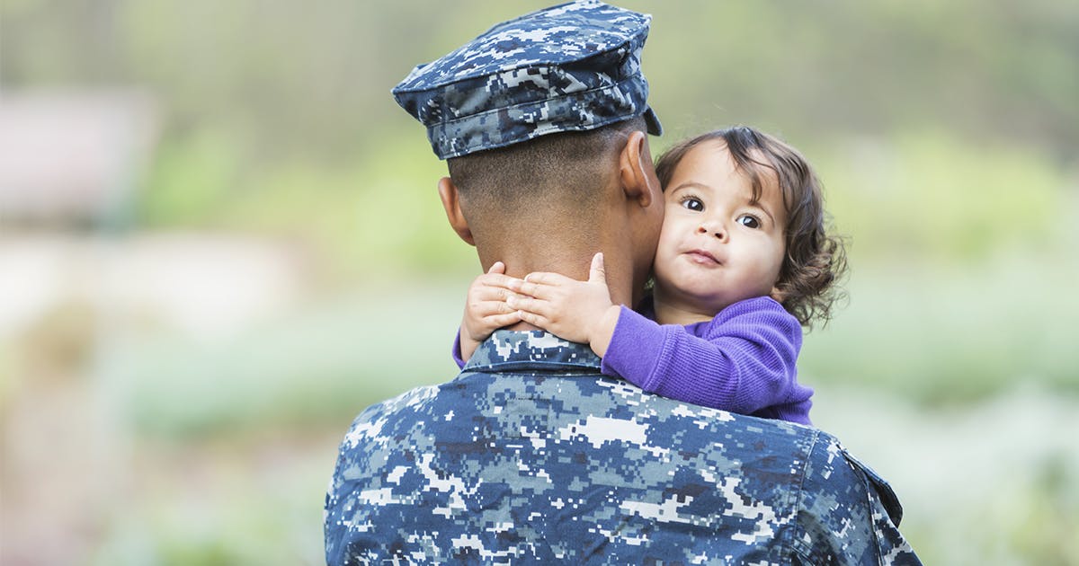 A little girl in a purple shirt wraps her arms around a man in military fatigues, pressing her cheek to his. 