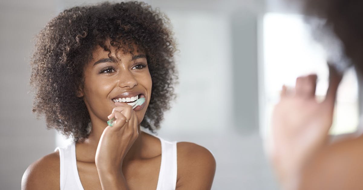 A woman with curly hair wearing a white tank top smiles into the mirror while brushing her teeth. 