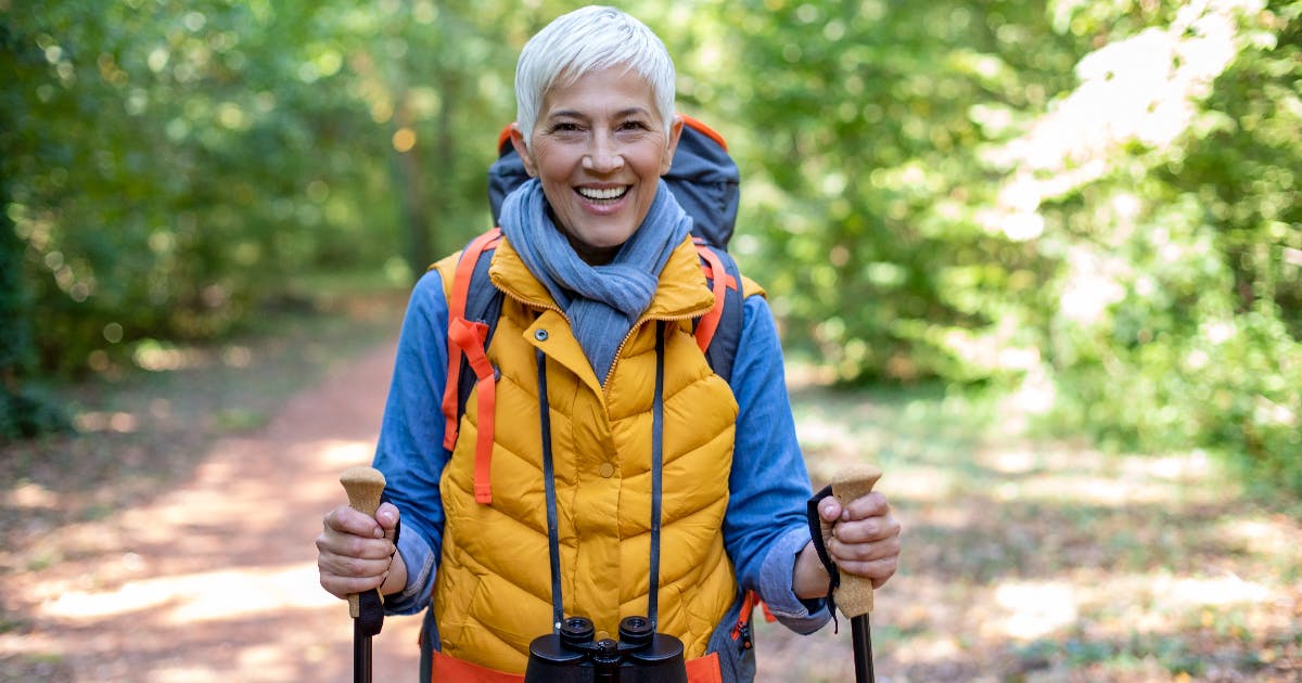 An older woman with hiking poles stands on a trail, smiling.