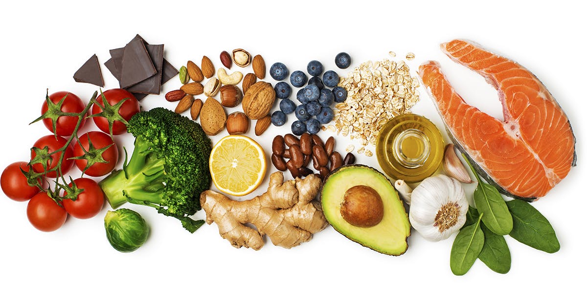 An assortment of heart healthy foods, including tomatoes, chocolate, broccoli, Brussels sprouts, nuts, lemon, ginger, blueberries, beans, ginger, avocado, oatmeal, olive oil, garlic, spinach and fish.