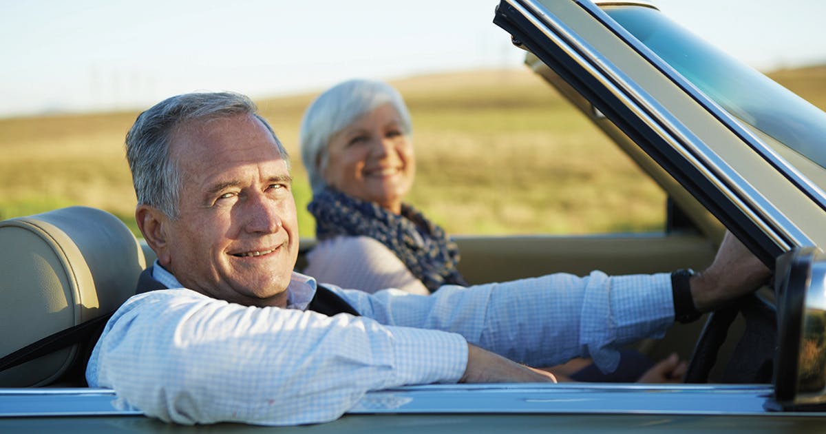 Two older adults riding in a car with the top down.