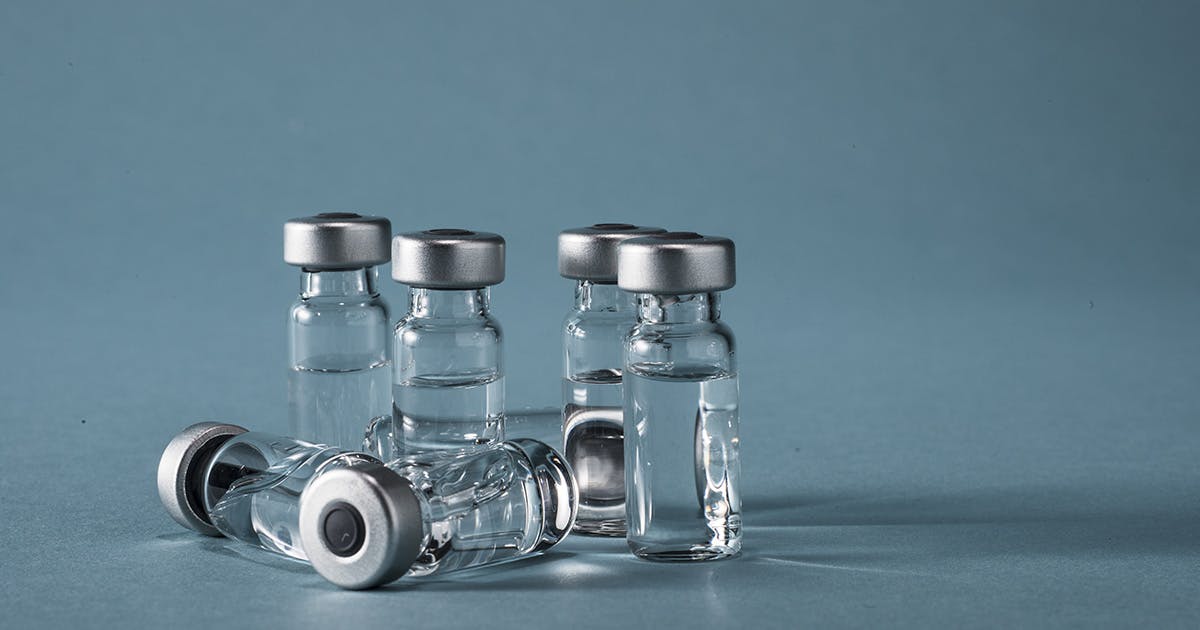 Several glass vials of clear vaccine liquid sit on a blue background. 