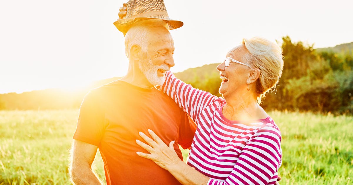 A smiling older couple hug as the woman playfully lifts the man's hat