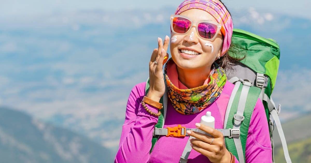 A woman in hiking gear and sunglasses spreads sunscreen on her face.