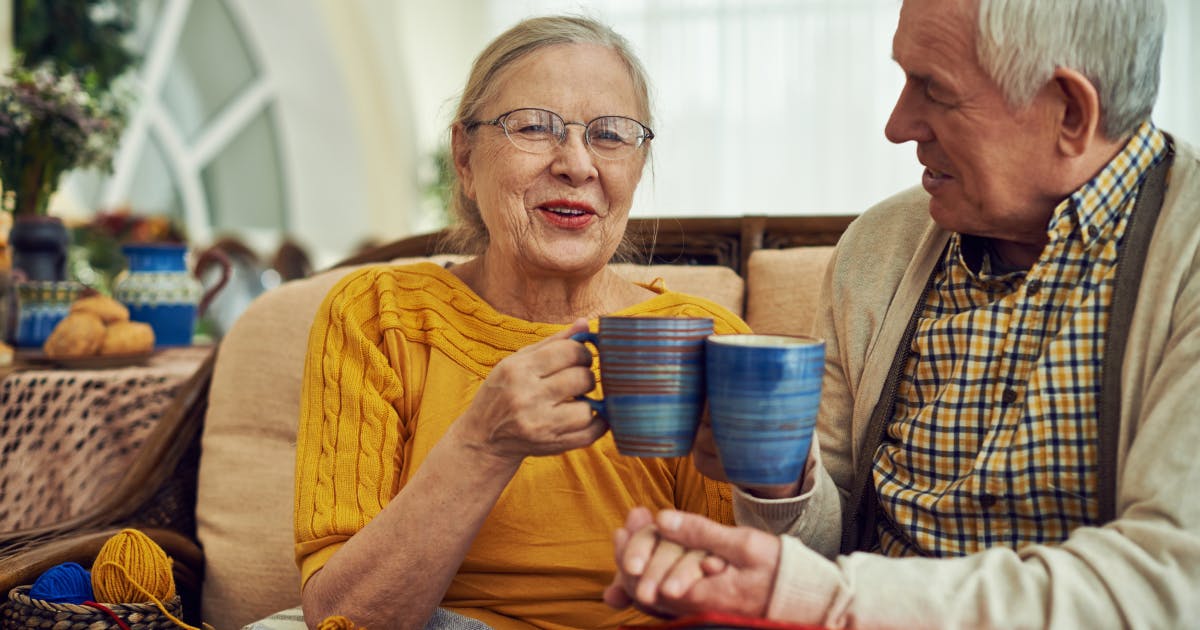 An older couple clinks their mugs together while holding hands on a couch.