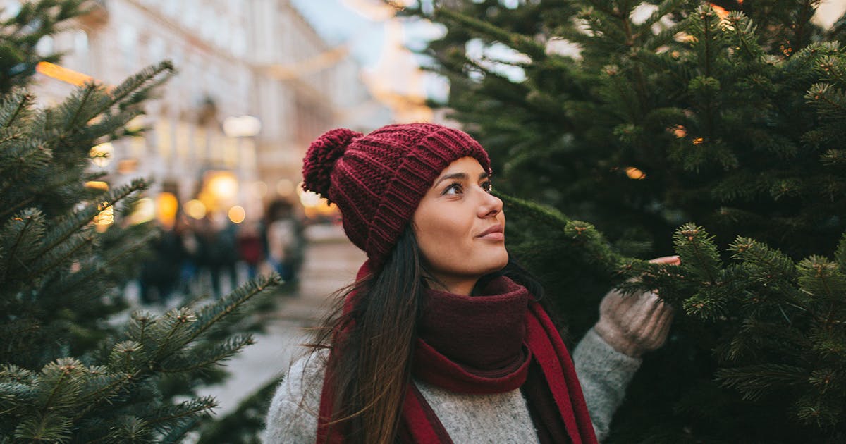 A young woman is standing between two trees in a Christmas tree lot.
