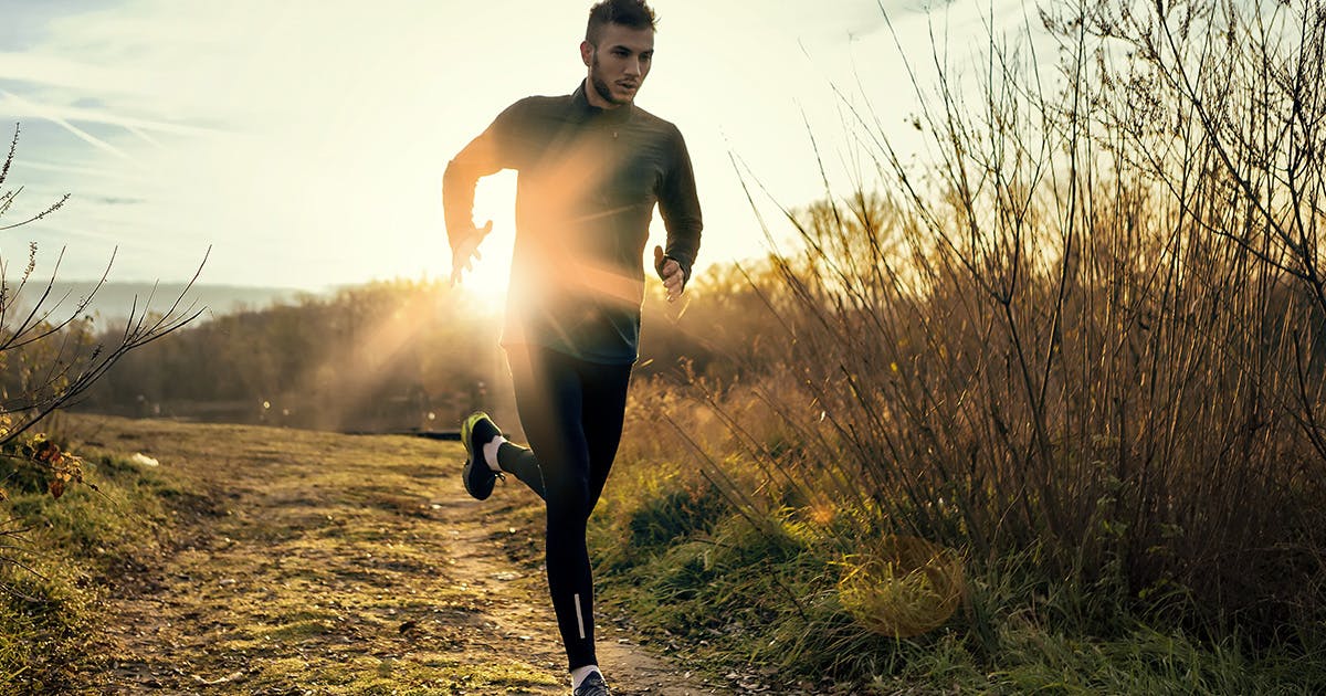 A young man runs along a trail with the sun rising behind him.