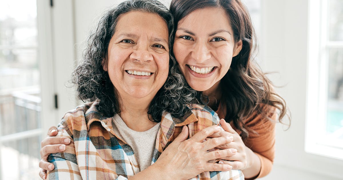 An adult woman standing close behind her mom with her hands on her mom's arms. 