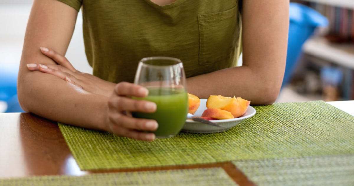 A woman holds a glass of green liquid in front of a plate of fruit.