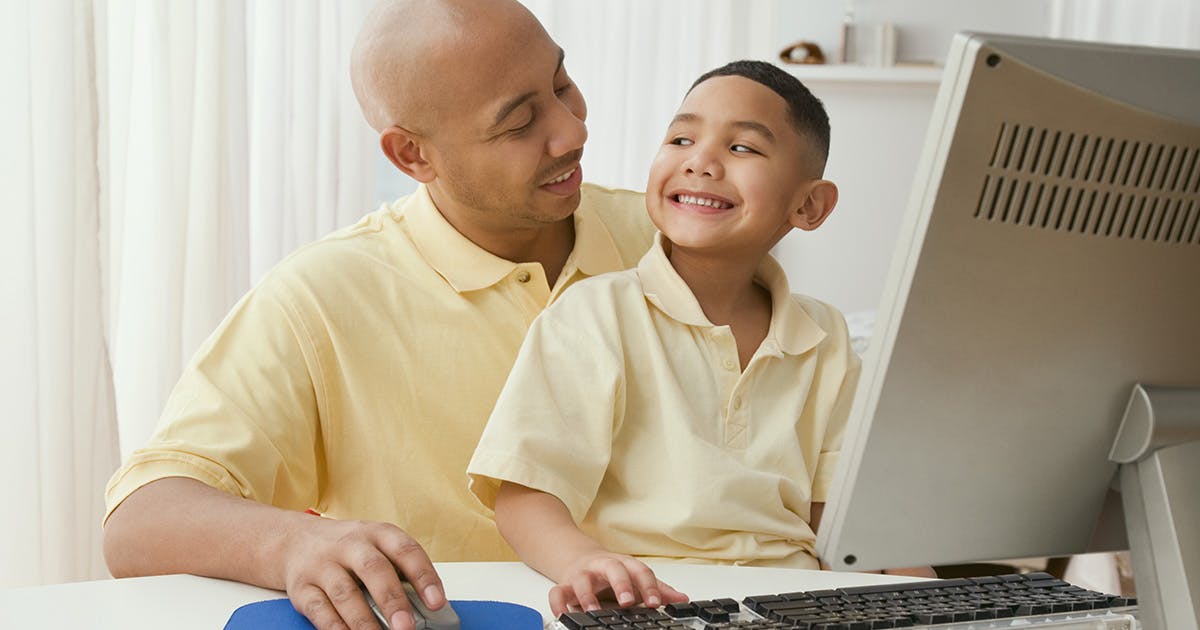 A father and son sit in front of a computer together and smile at each other. 